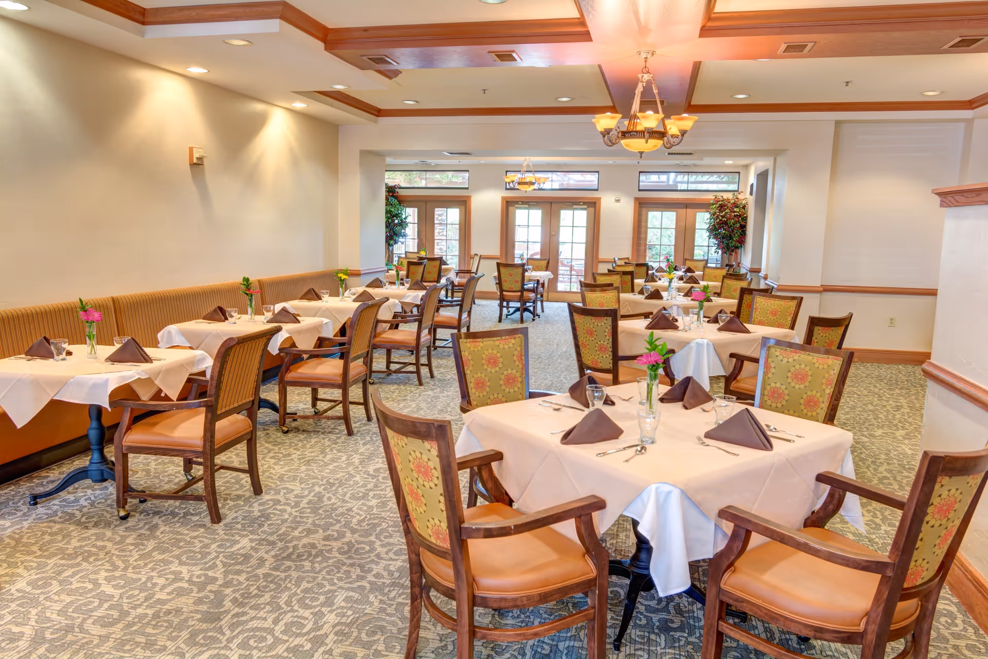 A bright and spacious dining room with multiple tables covered in white tablecloths and set with brown folded napkins, glassware, and silverware. The chairs have wooden frames with patterned upholstery. There are chandeliers hanging from the ceiling and large windows and glass doors letting in natural light. Small flower vases decorate each table.