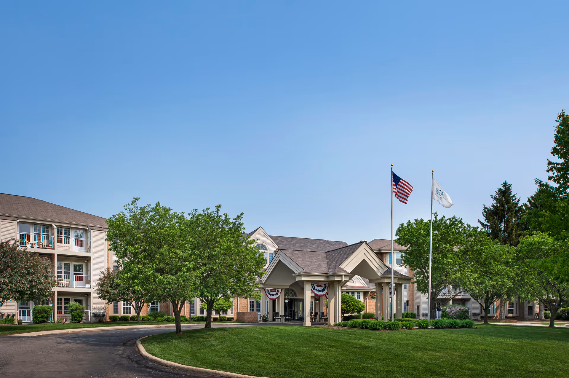 Exterior view of Baldwin House Senior Living Oakland showing a large, well-maintained building with multiple windows and balconies. The entrance features a covered driveway with columns, decorated with patriotic bunting. Two flagpoles stand in front, one with the American flag and the other with a white flag. The surrounding area has green grass, trees, and clear blue sky.