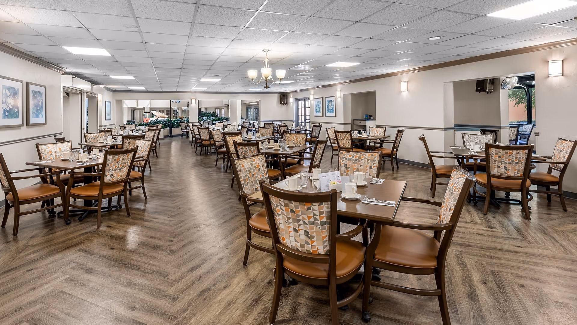 A spacious dining room with multiple wooden tables and chairs arranged neatly. Each table is set with white cups, saucers, and utensils. The room has a wood-patterned floor, light-colored walls with framed artwork, and ceiling lights providing bright illumination. There are large mirrors on the far wall and windows letting in natural light.
