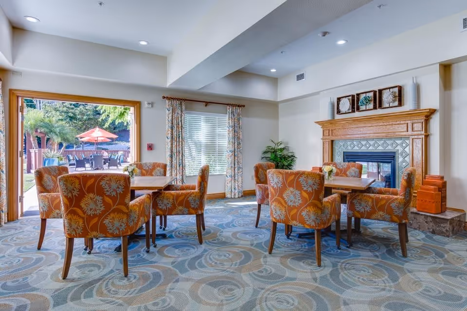 A cozy indoor seating area with two wooden tables surrounded by orange floral upholstered chairs. The room features a fireplace with decorative tiles and wooden mantel, patterned curtains on the windows, and a large open doorway leading to an outdoor patio with tables, chairs, and an orange umbrella. The carpet has a blue and beige swirling pattern.