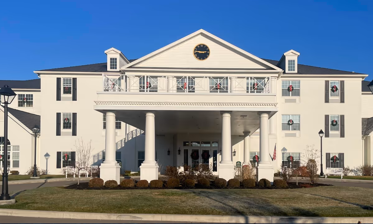 Front exterior view of a large white senior living facility building with a covered entrance supported by four large columns. The building has multiple windows decorated with holiday wreaths and a clock at the top center of the facade. There are lamp posts and landscaped bushes in front of the building under a clear blue sky.