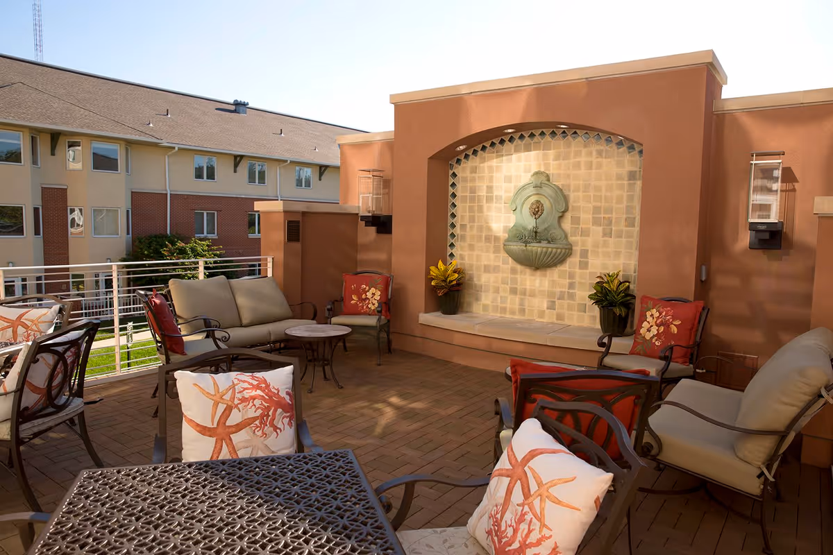 Outdoor patio area with cushioned chairs and a small table. The patio has a decorative wall fountain with plants on either side. The background shows a multi-story building with windows and a sloped roof.