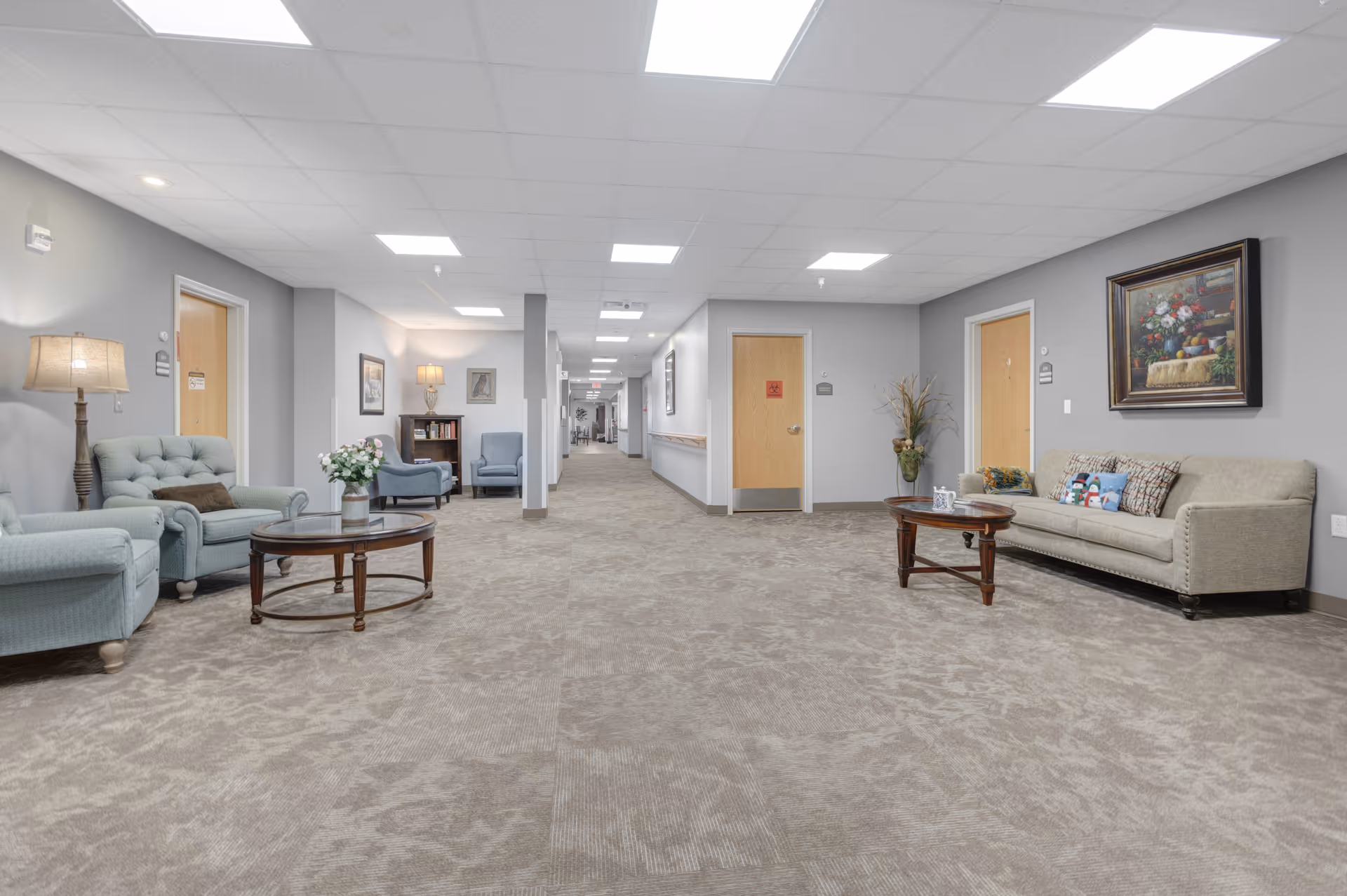 A spacious senior living facility hallway with beige carpet and gray walls. The hallway features comfortable seating areas on both sides, including armchairs, sofas, and wooden coffee tables with decorative items like flowers and mugs. The ceiling has recessed lighting, and there are several wooden doors along the hallway. A framed painting and a vase with plants decorate the walls.