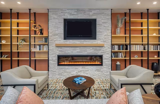A cozy living room area with two beige armchairs facing a modern electric fireplace set in a white stone wall. Above the fireplace is a mounted flat-screen TV. On either side of the fireplace are built-in wooden shelves with books and decorative items. A round wooden coffee table with books on it sits in front of the chairs on a patterned rug.
