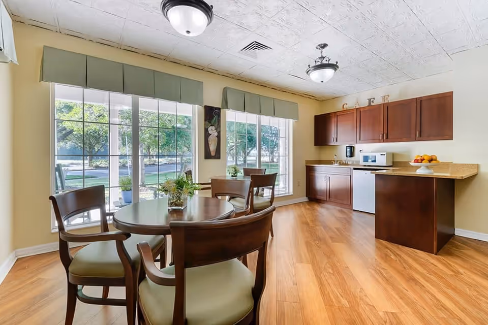 A bright and cozy cafe area with large windows showing greenery outside. The room features a round wooden table with four chairs, a small kitchenette with dark wood cabinets, a microwave, a mini fridge, a sink, and a countertop with a bowl of fruit. The ceiling has decorative tiles and two light fixtures.