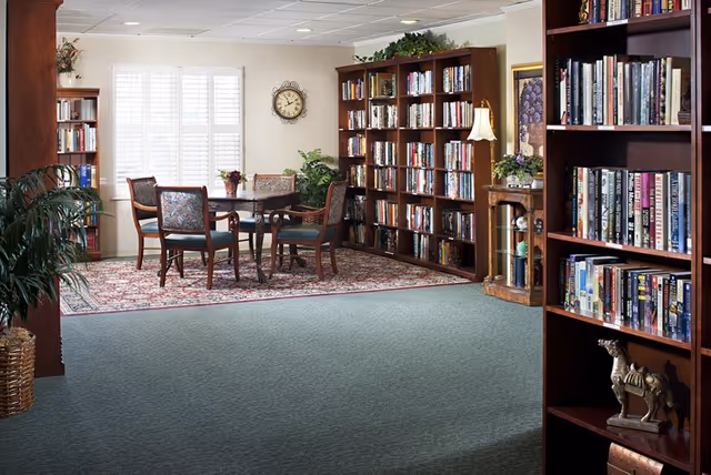 A cozy senior living facility common area with bookshelves filled with books, a round table with four chairs on a patterned rug, a wall clock, potted plants, and a small wooden cabinet with a lamp and decorative items.