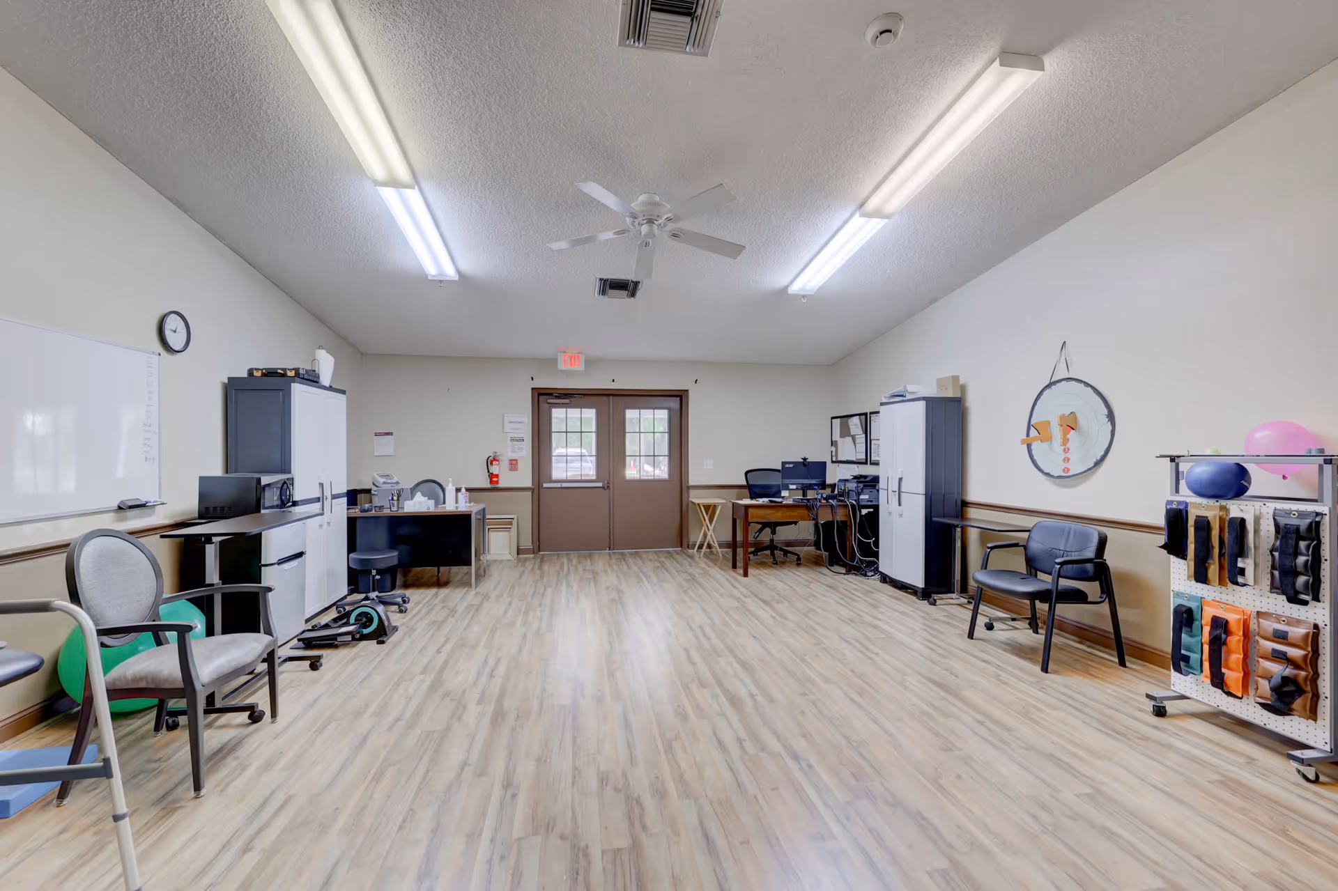 A spacious room with light wood flooring and white walls, featuring two desks with office chairs, storage cabinets, a whiteboard, and various exercise and therapy equipment including weights and exercise balls. The room has fluorescent ceiling lights, a ceiling fan, and double doors with windows at the far end.