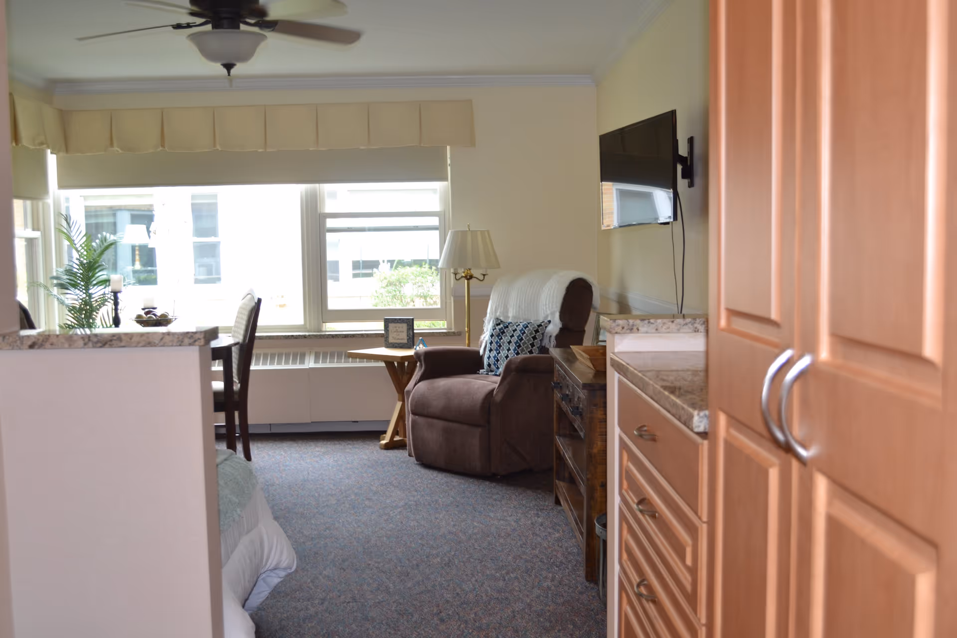 Interior view of a senior living facility room at Gilbert Residence showing a cozy armchair with a pillow and a white throw blanket, a small wooden side table with a lamp and a framed picture, a mounted flat-screen TV on the wall, a dining table with chairs near large windows with blinds, and wooden cabinetry in the foreground.