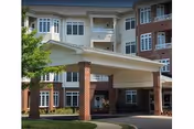 Entrance of a multi-story senior living facility with a covered drop-off area supported by brick columns. The building has large windows and a mix of brick and light-colored siding. There is a tree and some landscaping visible near the entrance.