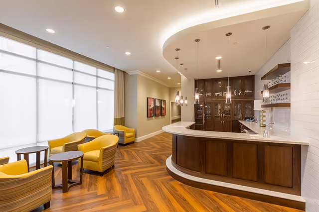 A bright and modern lounge area with yellow armchairs and small round tables on a wood-patterned floor. To the right is a curved wooden bar with a marble countertop, pendant lights hanging above, and glass cabinets behind the bar. Large windows with white blinds allow natural light to fill the space.