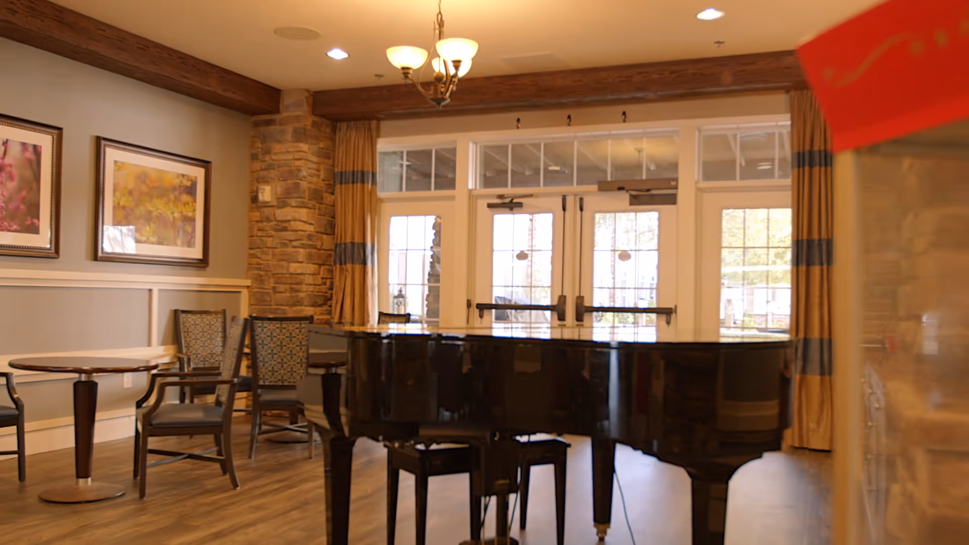 Sunlit common room with a black grand piano, seating, small tables, framed artwork, and glass double doors.