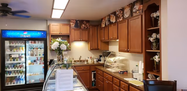 Small kitchenette-style snack area with wooden cabinets, a Pepsi beverage cooler, glass display cases, and a counter with a sink and flowers.
