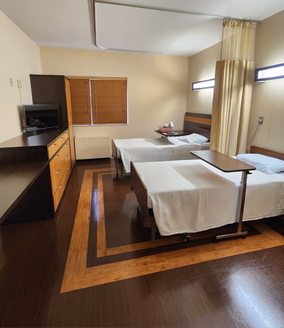 A room with two single beds covered in white sheets, each with a small overbed table. The room has wooden flooring with a decorative border, a window with closed wooden blinds, a dresser with multiple drawers, and a small TV on top. There is a privacy curtain between the beds and wall-mounted lights above each bed.