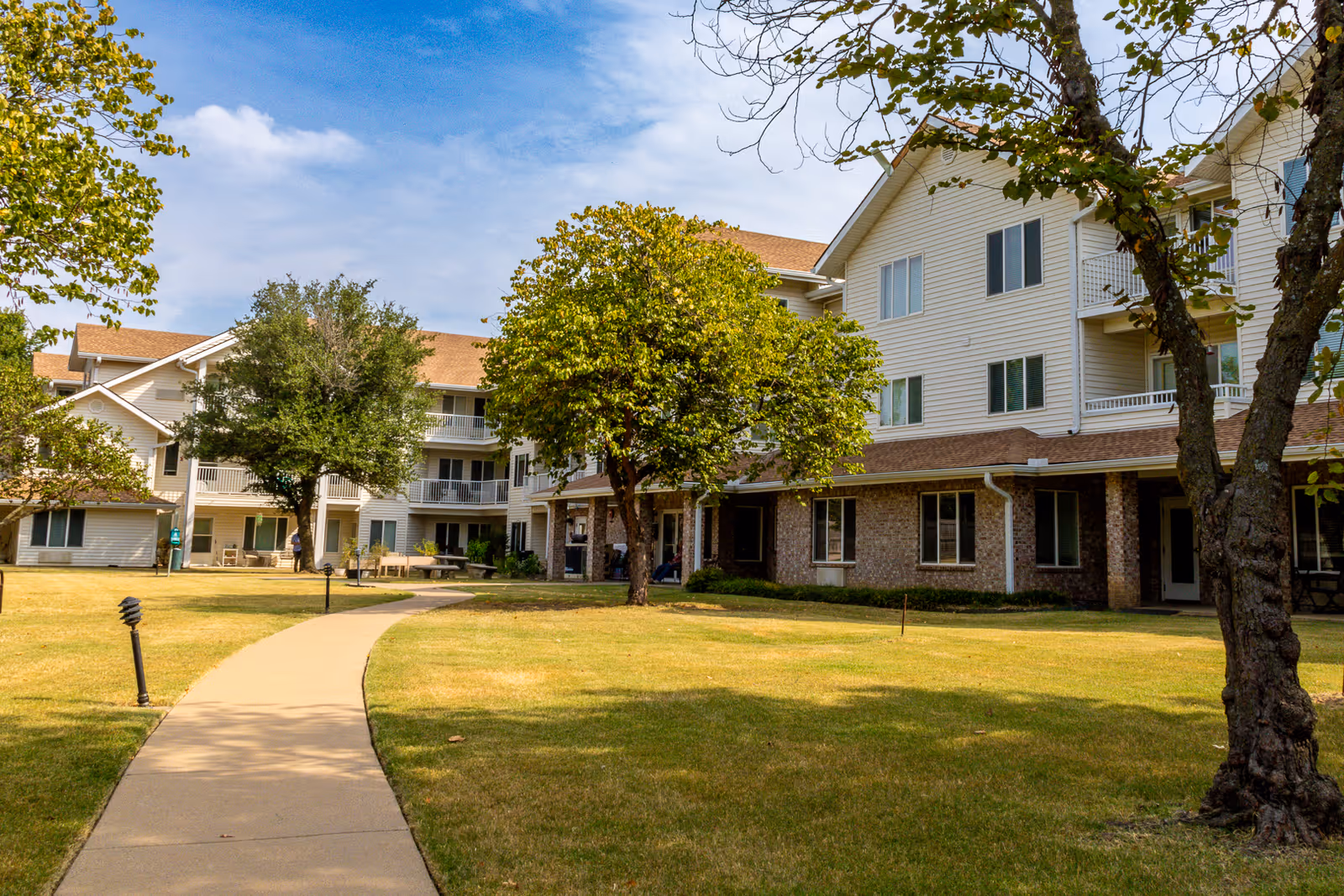 Exterior view of a multi-story senior living facility with beige siding and brick accents, surrounded by a well-maintained lawn with trees and a paved walkway leading towards the building under a partly cloudy sky.