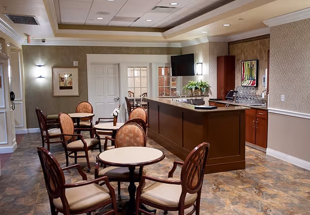A cozy interior room with multiple round tables and cushioned chairs arranged for seating. There is a wooden counter with a granite top, a mounted flat-screen TV, and cabinetry with a sink. The walls are decorated with framed artwork and have a textured wallpaper. The floor has a patterned tile design, and the ceiling features recessed lighting.
