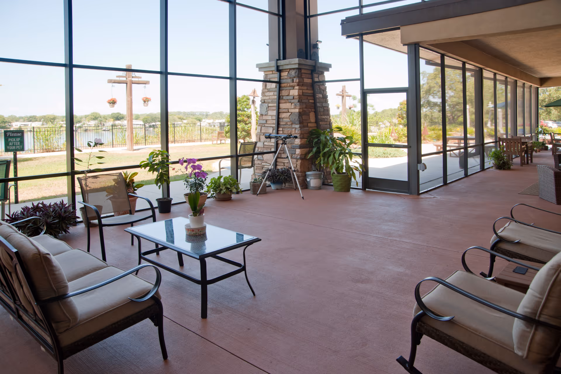A spacious screened-in patio area with cushioned chairs, a glass-top coffee table with a potted orchid, several potted plants, and a stone fireplace. Large windows provide a view of an outdoor garden and water body beyond.
