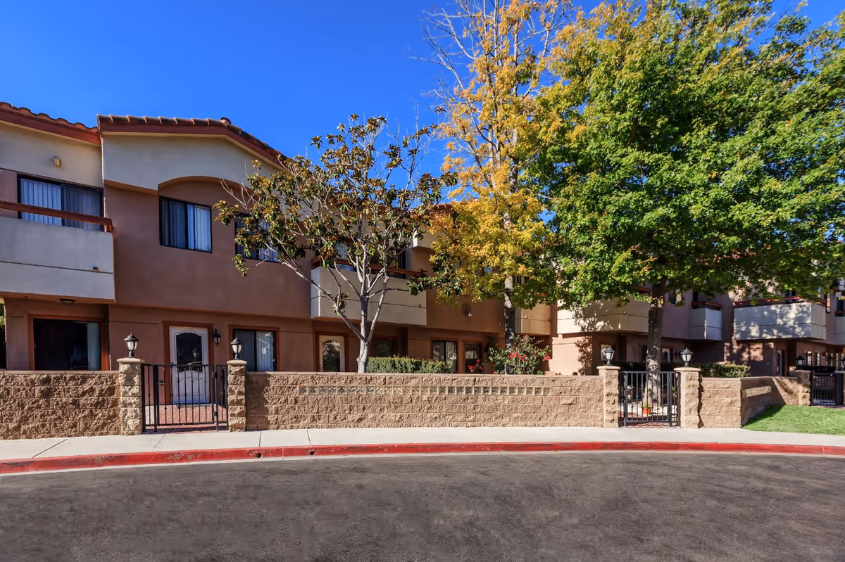 Exterior view of a two-story residential building with beige and brown walls, balconies, and several windows. There are trees with green and yellow leaves in front of the building, a low brick wall with black metal gates, and a sidewalk along a curved road under a clear blue sky.