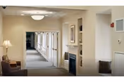 A well-lit interior hallway in a senior living facility with beige walls and carpeted floors. The hallway features a handrail along the left side, a small seating area with a lamp and chair on the left, and a built-in fireplace with framed artwork above it on the right. The space appears clean and welcoming.