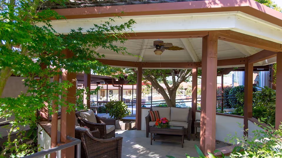 Outdoor covered seating area with cushioned wicker chairs and a sofa arranged around a coffee table, surrounded by greenery and trees, with a ceiling fan overhead.