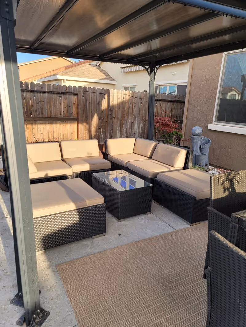 Covered backyard patio with rattan sectional seating, cushioned ottomans and a glass-top coffee table under a pergola.