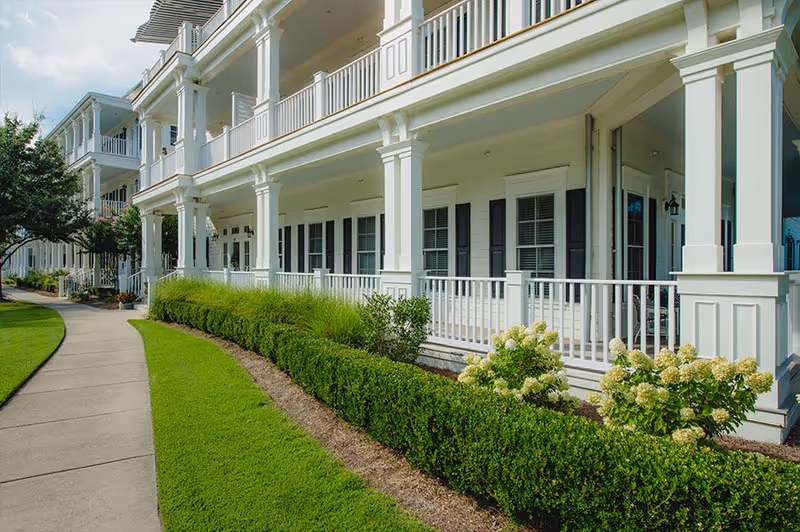 Exterior view of a white multi-story building with a covered porch, white railings, and black shutters on the windows. The building is surrounded by neatly trimmed bushes, flowering plants, and a well-maintained lawn with a curved concrete walkway.