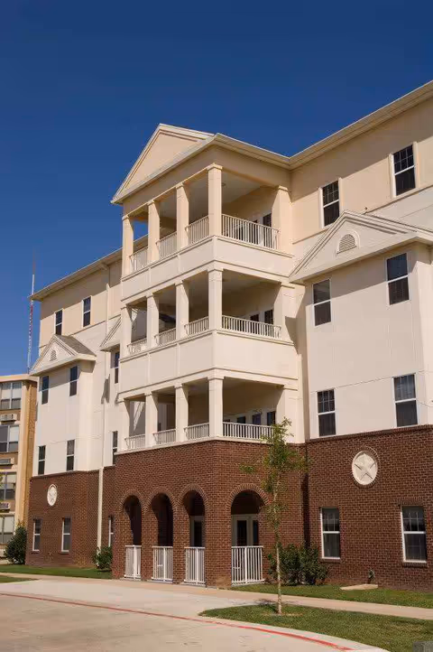 Exterior view of a multi-story senior living facility building with beige and brown brick facade, featuring balconies with white railings and columns under a clear blue sky.