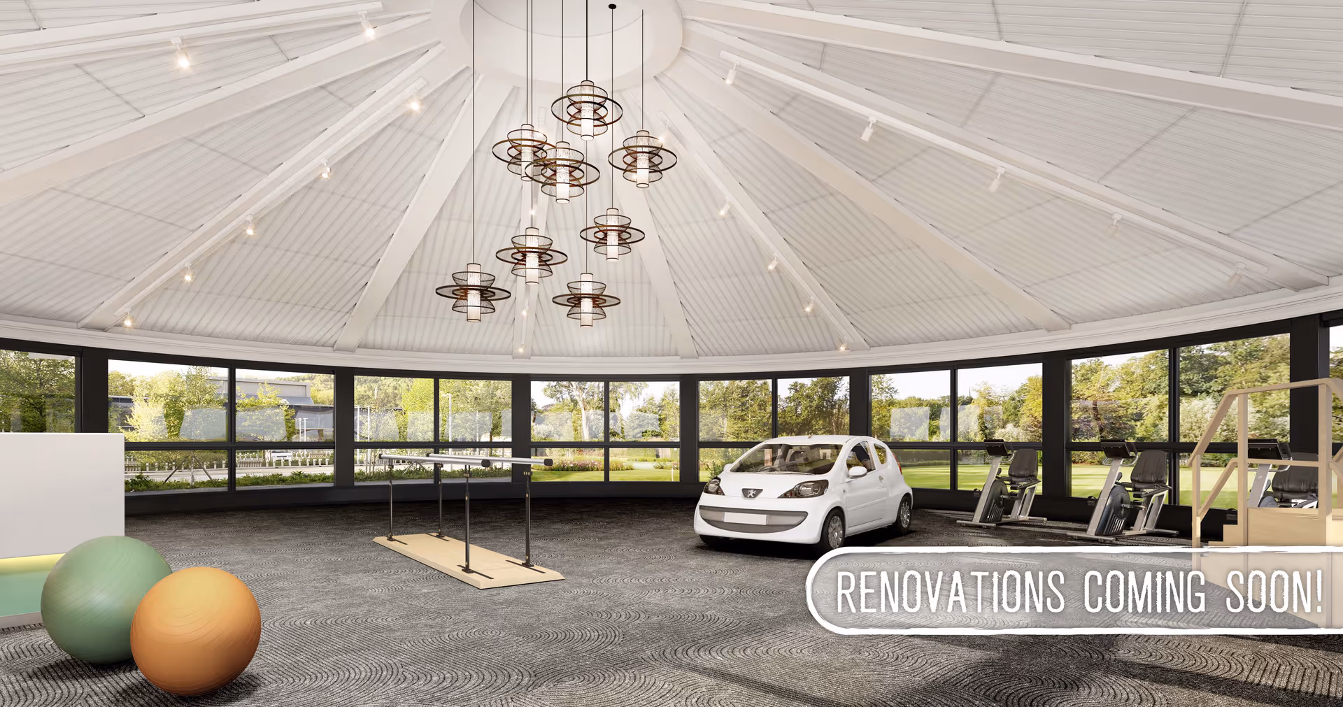A spacious rehabilitation room with large windows showing greenery outside. Inside, there are exercise equipment including stationary bikes, a small white car for mobility training, parallel bars for walking practice, and two large exercise balls. The ceiling is high and white with modern hanging light fixtures. Text on the image reads 'RENOVATIONS COMING SOON!'