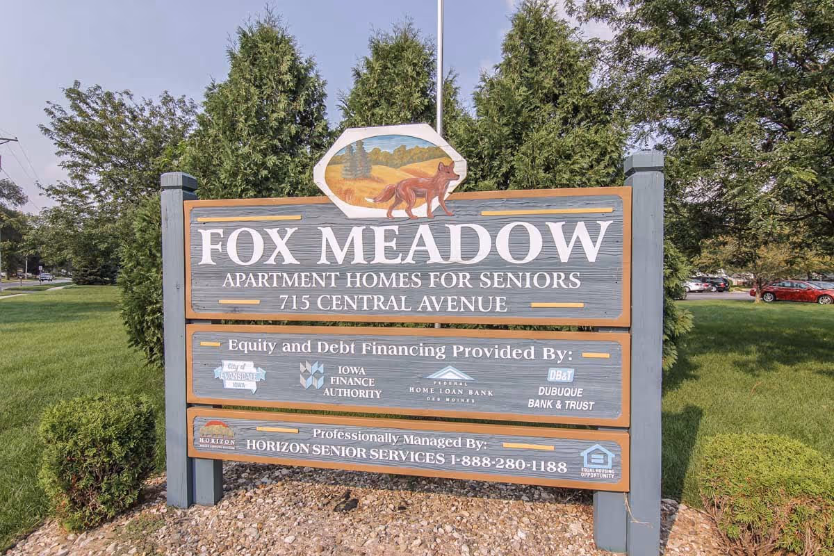 Wooden entrance sign reading 'Fox Meadow Apartment Homes for Seniors' on a grassy lawn with trees and parked cars in the background.