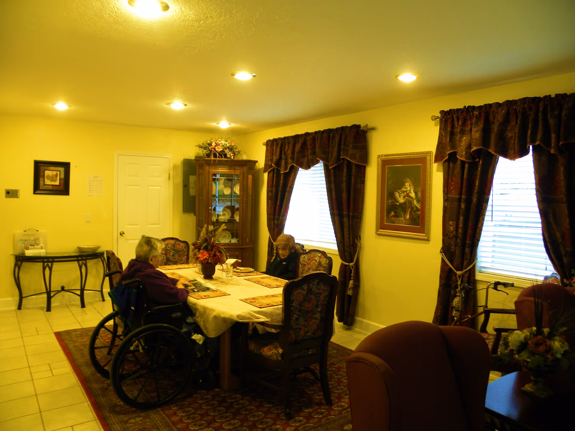 A cozy dining room with a rectangular table covered with a tablecloth and placemats. Two elderly women are seated at the table, one in a wheelchair. The room has floral curtains on the windows, a framed painting on the wall, a wooden cabinet with decorative plates, and a floral centerpiece on the table. The floor is tiled with a patterned rug under the table.