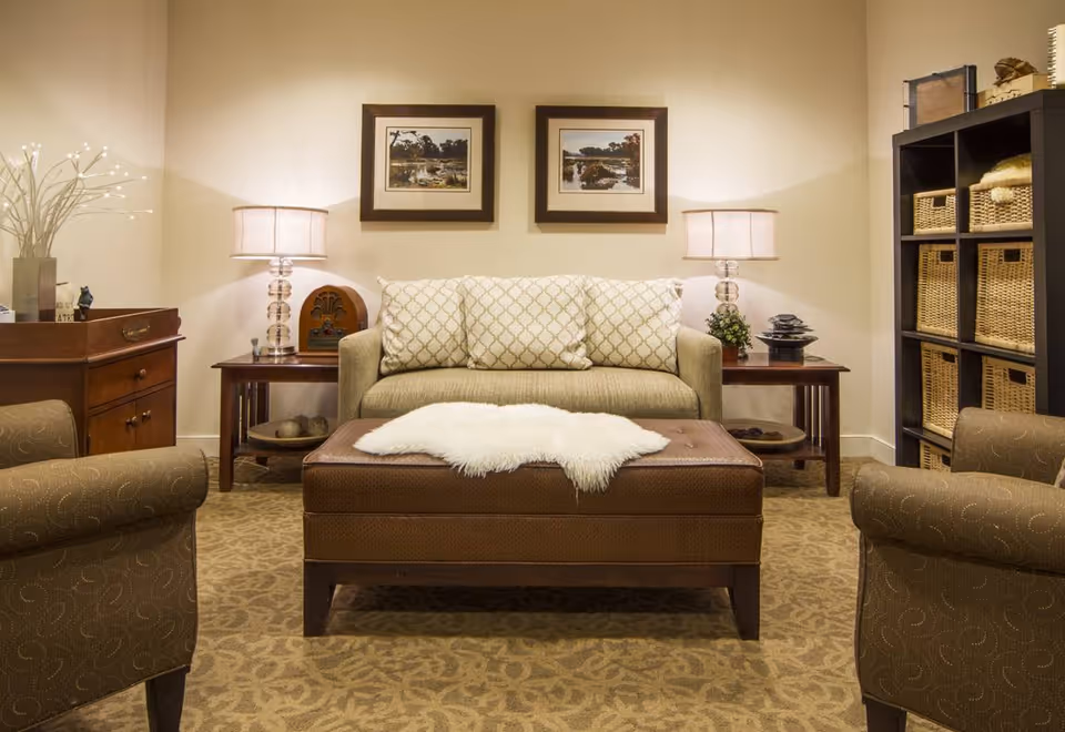 A cozy living room with a beige couch adorned with three patterned cushions, two matching armchairs, and a brown ottoman with a white fur throw on top. The room features two side tables with lamps, framed landscape artwork on the wall, a wooden cabinet on the left, and a black shelving unit with wicker baskets on the right.