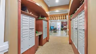 Interior view of a hallway with multiple mailboxes on both sides, leading to a common area with tables and chairs in the background. The hallway has warm lighting and decorative plants on top of the mailbox units.