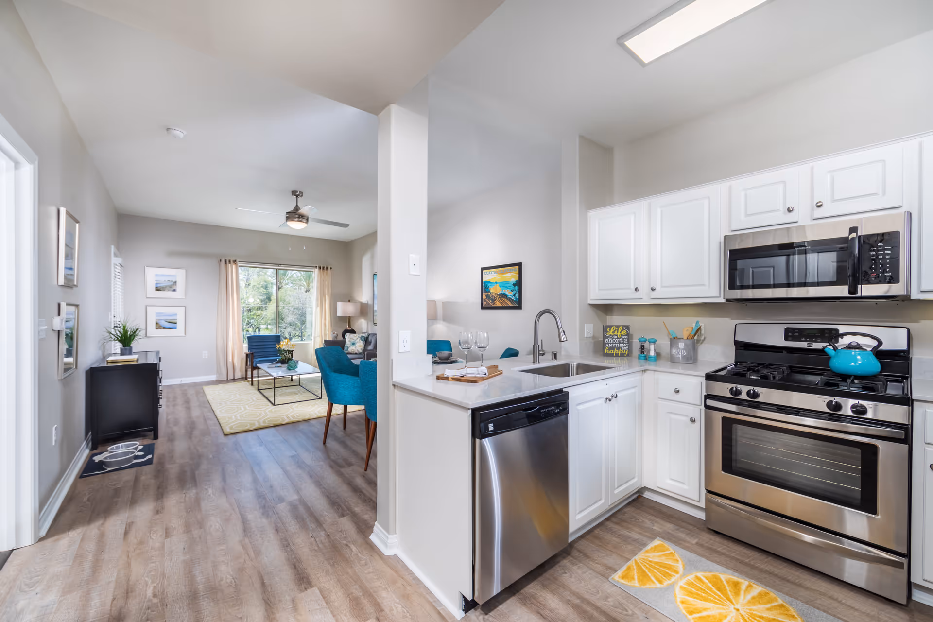 Bright and modern open-concept kitchen and living room area. The kitchen features white cabinets, stainless steel appliances including a dishwasher, oven, and microwave, and a sink with a faucet. A small rug with lemon slices is placed in front of the oven. The living room has a ceiling fan, a large window with beige curtains, a yellow patterned rug, blue chairs, a coffee table, and framed pictures on the walls. The flooring is wood-style laminate throughout.
