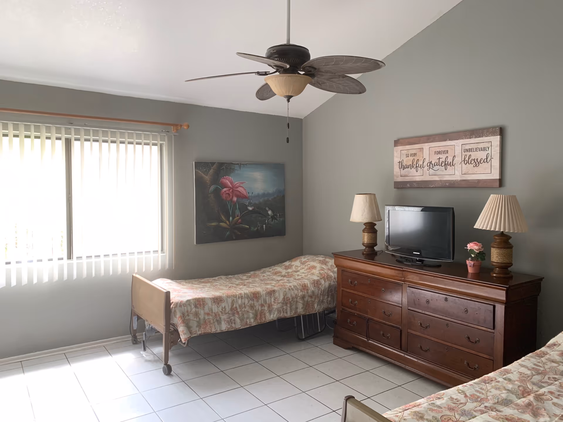 A simple bedroom with two single beds covered in floral bedspreads, a wooden dresser with a small TV, two table lamps, and a small potted plant. The walls are painted gray and decorated with two framed artworks. A ceiling fan with leaf-shaped blades hangs from the ceiling, and a window with vertical blinds allows natural light into the room.
