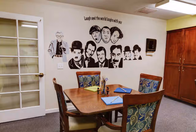 A small meeting or activity room with a wooden table and four chairs with patterned upholstery. On the table are notebooks, pencils, and paintbrushes. The wall features black and white caricature-style portraits of famous comedians with the phrase 'Laugh and the world laughs with you.' There is a glass-paneled door on the left and wooden cabinets on the right.