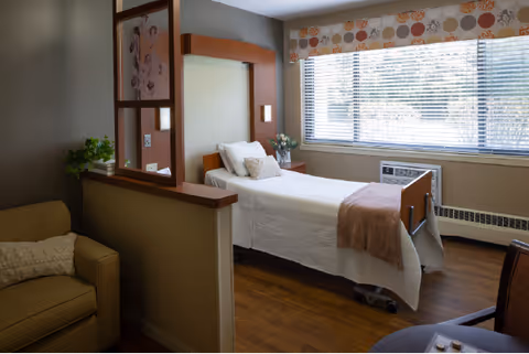 Furnished bedroom with a single adjustable bed, bedside table, large window with blinds, and a small seating area.