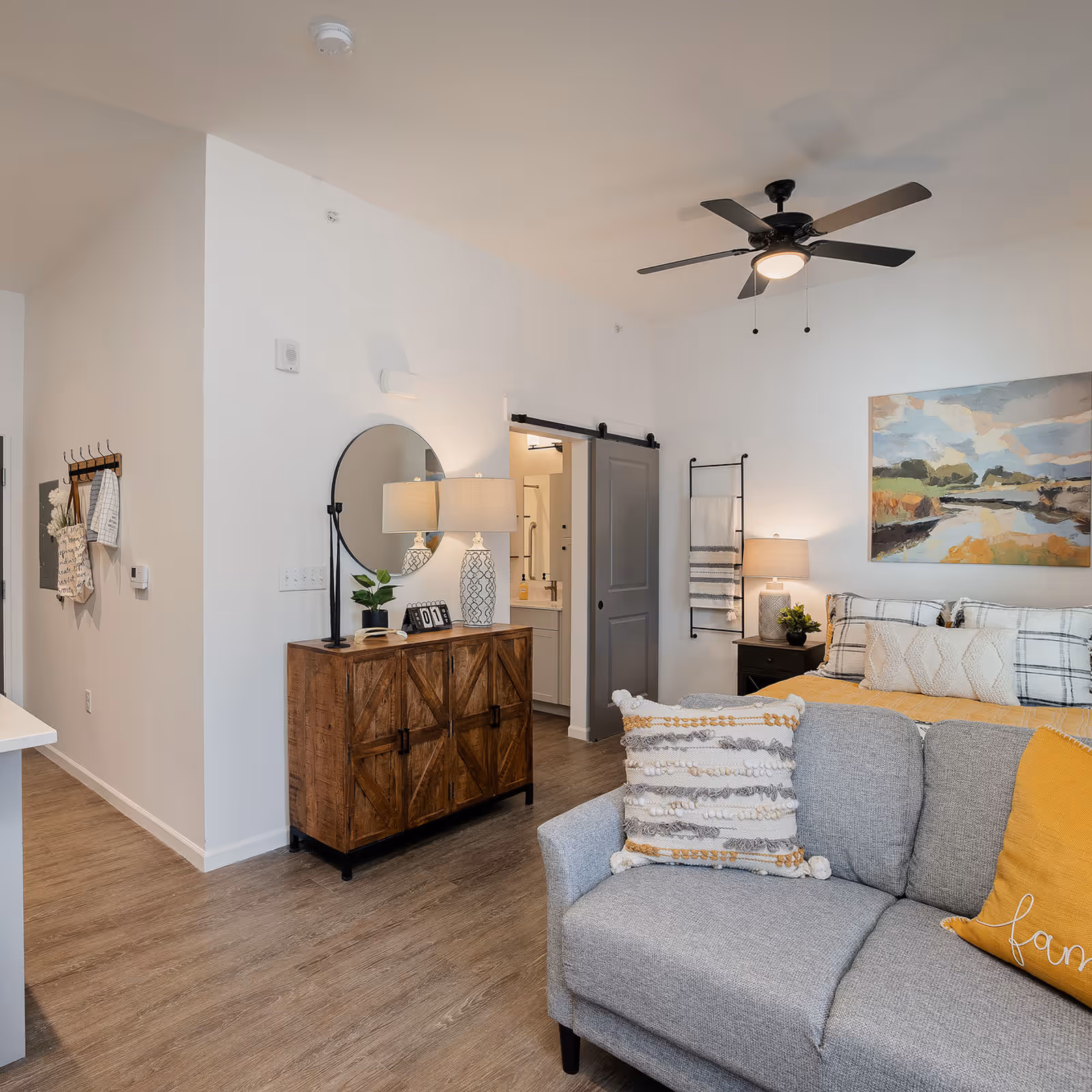 A cozy senior living apartment interior featuring a gray couch with decorative pillows in the foreground, a wooden cabinet with a round mirror and two lamps on top, a bed with plaid bedding and a yellow throw blanket, a bedside table with a lamp and plant, a ceiling fan, and a sliding barn door leading to a bathroom.