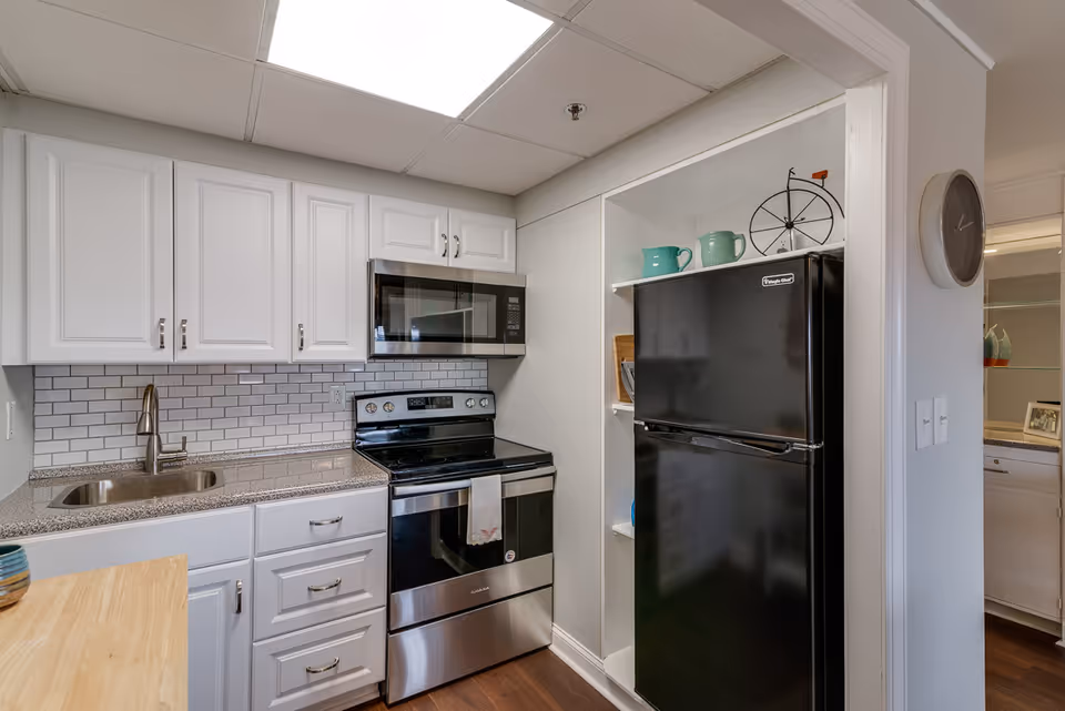 A compact kitchen with white cabinets, a stainless steel microwave and stove, a black refrigerator, a small sink with a modern faucet, and a wooden countertop. There are decorative items on the shelf above the refrigerator and a wall clock on the adjacent wall.