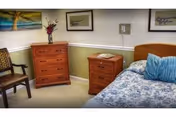 Small bedroom with a bed dressed in blue patterned bedding, a wooden nightstand and dresser, a chair, and framed artwork on the walls.