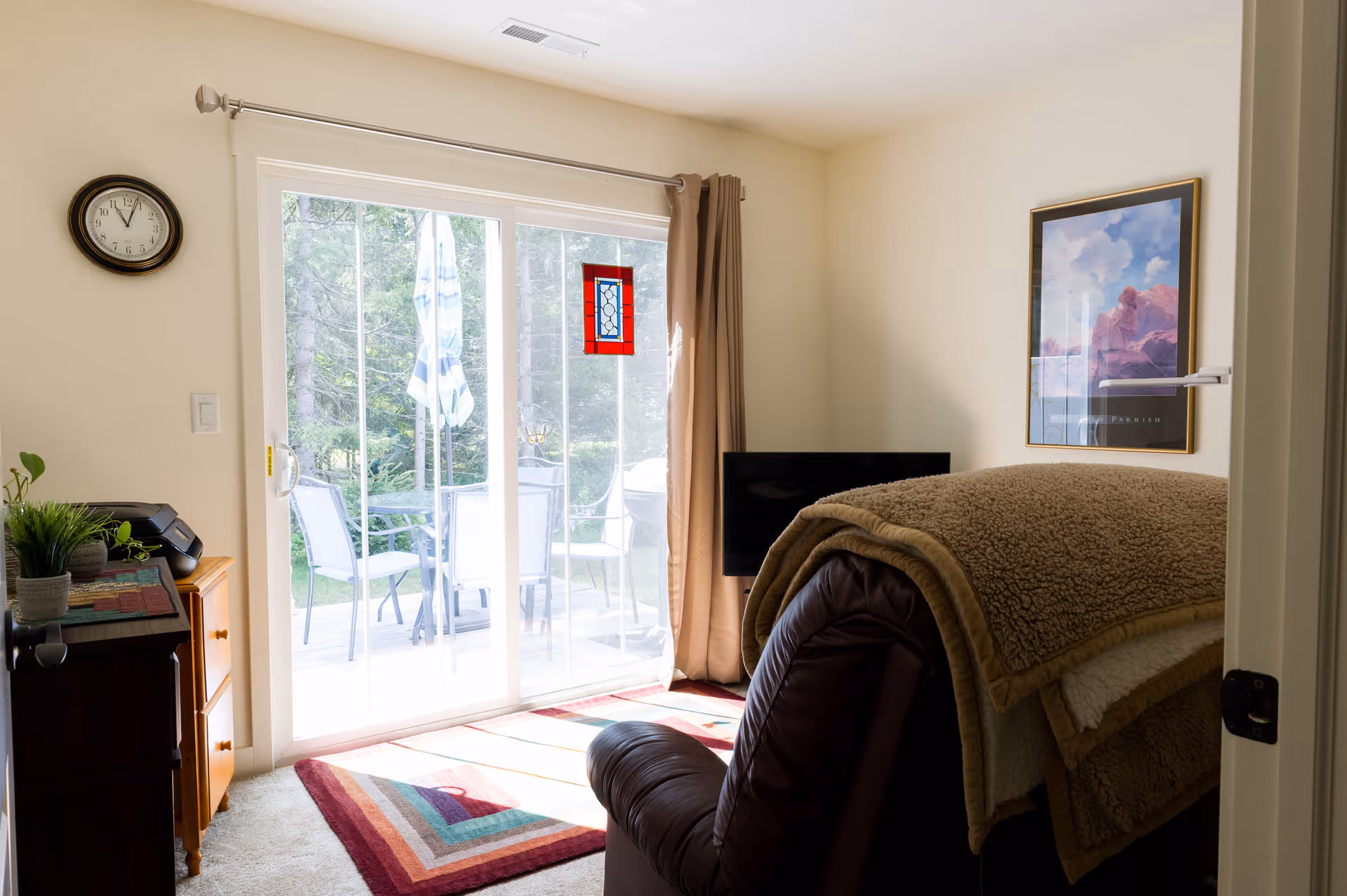 A cozy living room with a large brown recliner covered with a beige blanket facing a flat-screen TV. There is a colorful geometric rug on the carpeted floor and a sliding glass door leading to an outdoor patio with chairs and a table. A clock hangs on the wall to the left, and a framed picture is on the right wall. Light beige walls and curtains frame the sliding door.