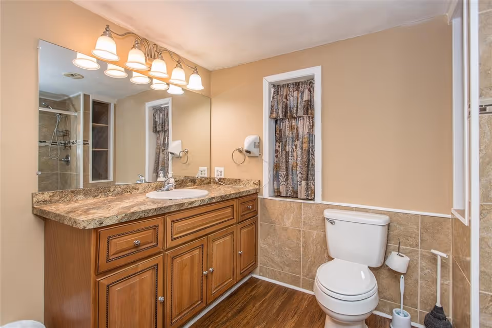 Well-lit bathroom with a wooden vanity and stone countertop with sink, a toilet, and a tiled shower seen in the mirror.