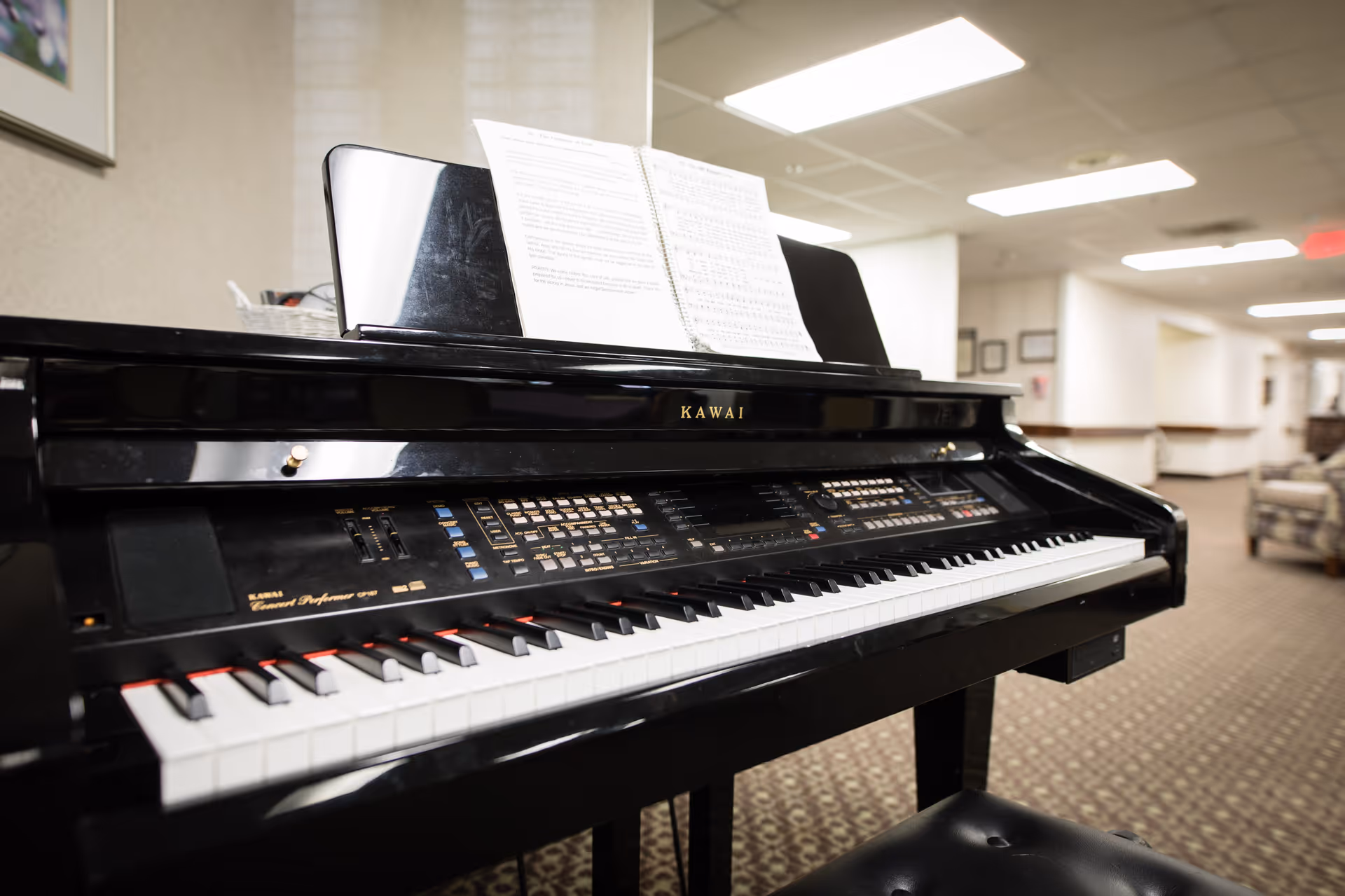 Black Kawai piano with sheet music on its stand in a carpeted common area/hallway of an assisted living facility.