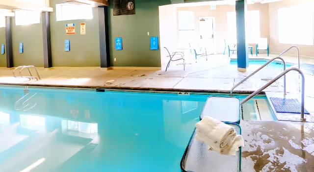 Indoor swimming pool area with handrails, lounge chairs, and folded towels on a poolside bench.