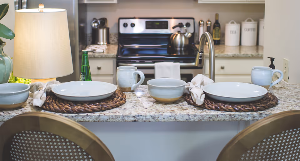 A kitchen counter set for two with ceramic bowls, plates, mugs, and cloth napkins on woven placemats. In the background, there is a stove, a lamp, and containers labeled Tea and Coffee on the countertop.