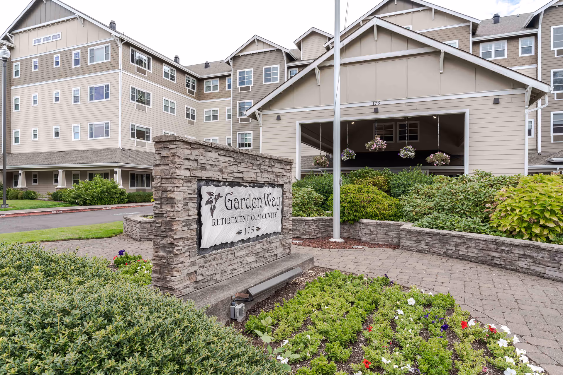 Exterior view of Garden Way Retirement Community building with a stone sign in the foreground displaying the community name and address. The building is multi-story with beige siding and white trim, surrounded by well-maintained landscaping including bushes and flowers.