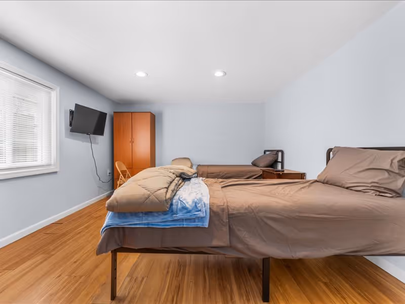 A simple bedroom with two single beds covered in brown bedding. There is a wooden wardrobe and a folding chair against the wall. A flat-screen TV is mounted on the wall near a window with blinds. The room has light blue walls and wooden flooring.