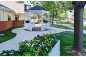A peaceful outdoor area at a senior living community featuring a white gazebo with hanging flower baskets, a curved concrete walkway, green grass, a large tree, and a flower bed with colorful blooms in the foreground.