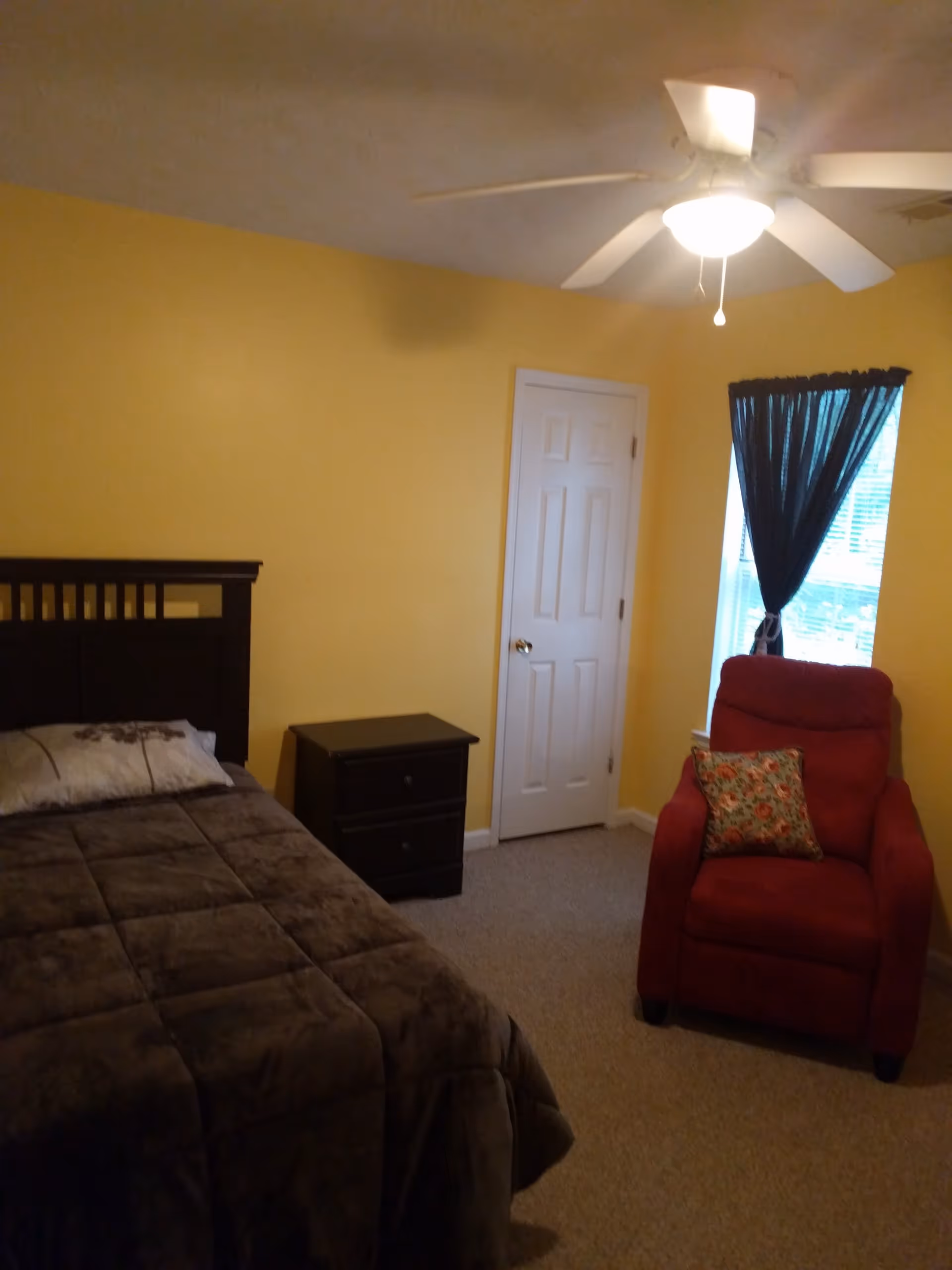 A bedroom with yellow walls featuring a bed with a dark brown headboard and a dark brown comforter, a matching nightstand with two drawers, a red armchair with a floral cushion, a window with dark curtains, a white door, and a ceiling fan with a light.