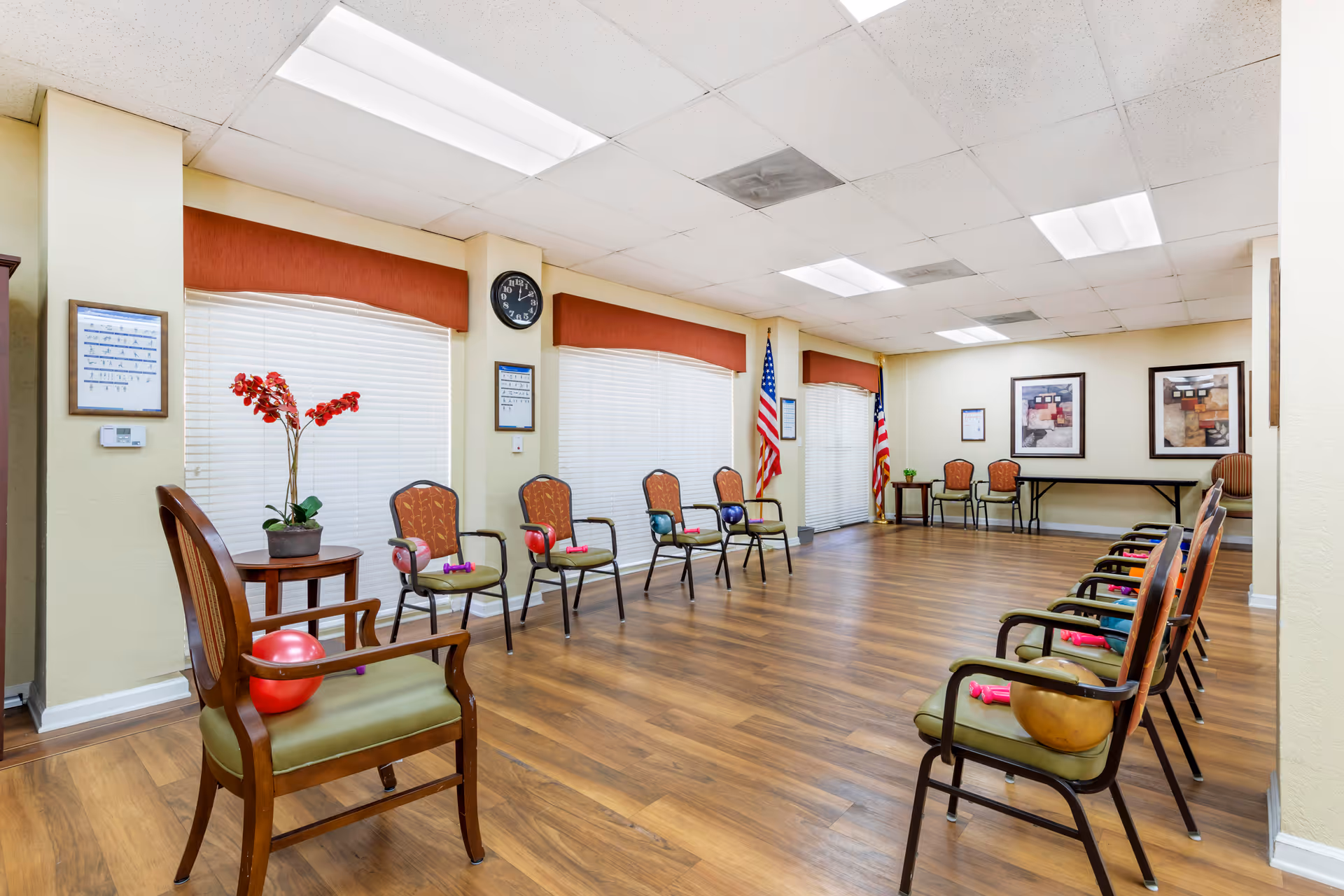 A spacious room with wooden flooring and a row of chairs arranged in a semi-circle. Each chair has a small exercise ball or dumbbells placed on or near it. The room has large windows with blinds and red valances, two American flags in the corner, framed artwork on the walls, and a clock above the windows. There is a small table with a potted plant and a side table with a red orchid flower arrangement.