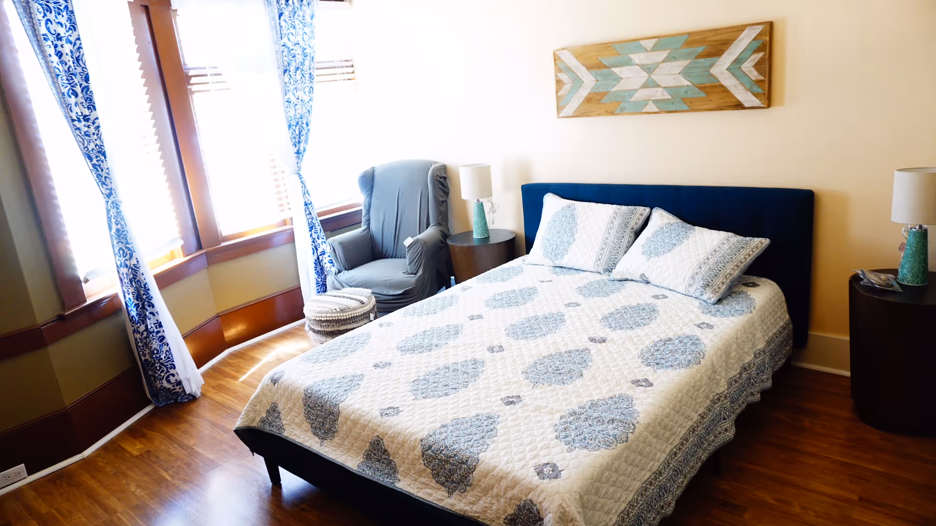 A bright bedroom with a bed covered in a white and blue patterned quilt, two matching pillows, a dark blue headboard, and wooden flooring. There is a gray armchair next to a round side table with a lamp, and blue and white patterned curtains hang on the windows. A decorative wooden wall art piece with geometric patterns is mounted above the bed.