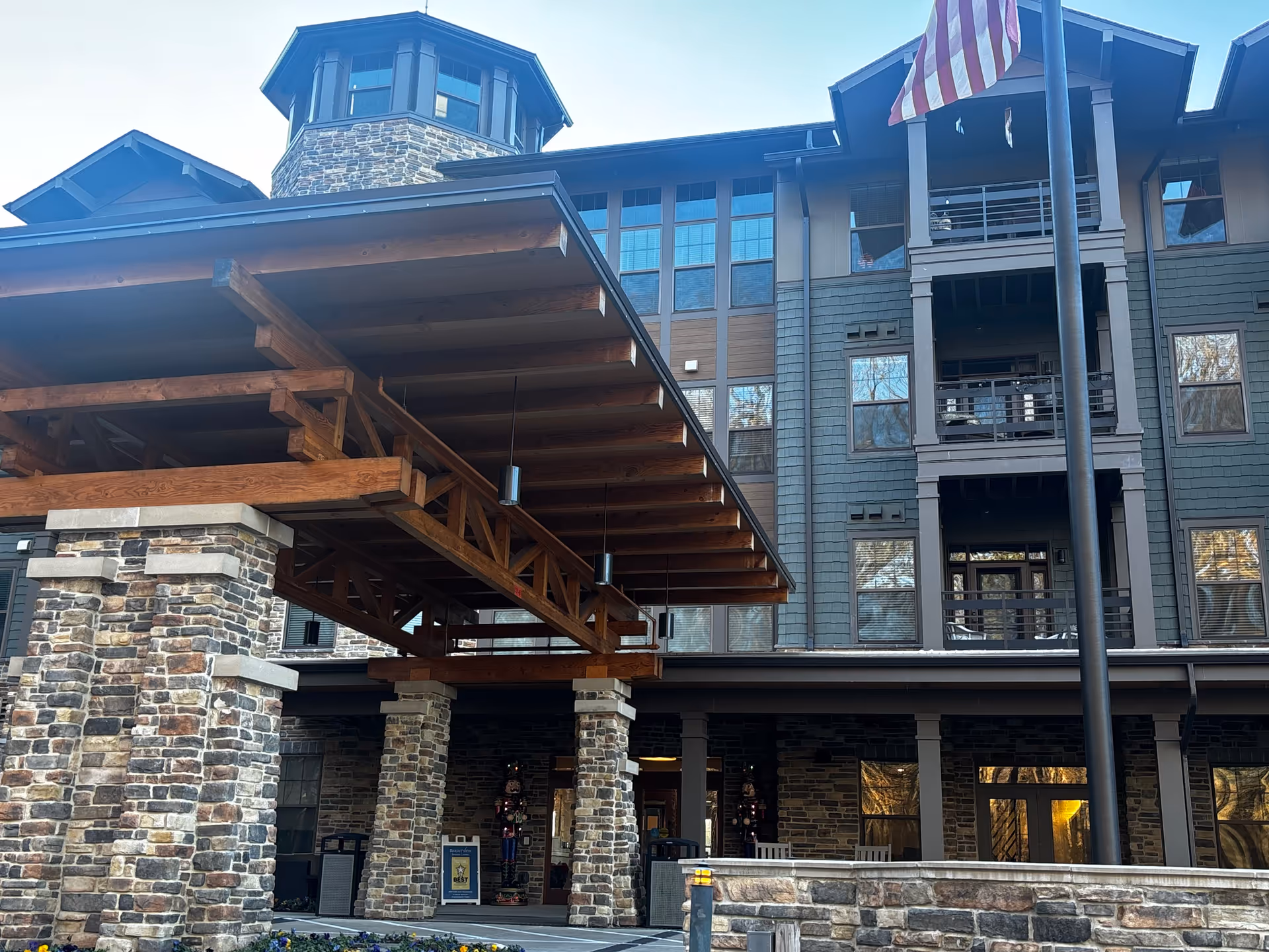 Front entrance of a multi-story senior living building with stone pillars, a large wooden canopy, and an American flag.
